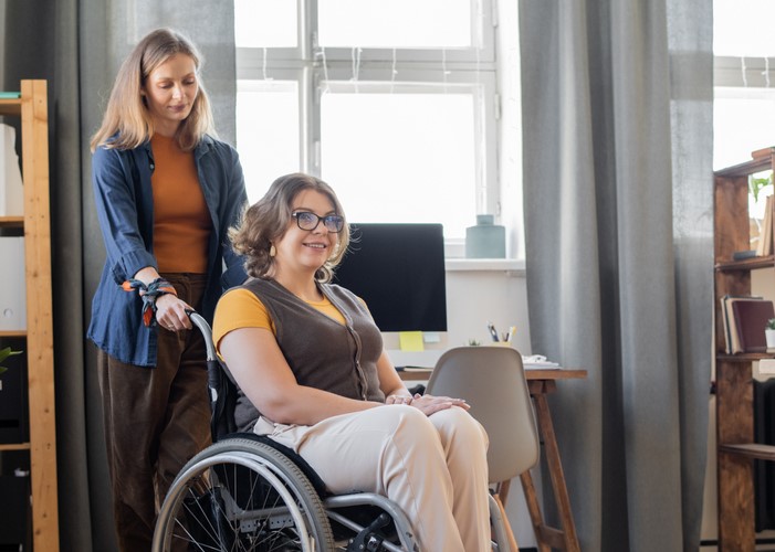 Young smiling woman in wheelchair looking forwards while her sister or friend standing behind and helping her with moving around