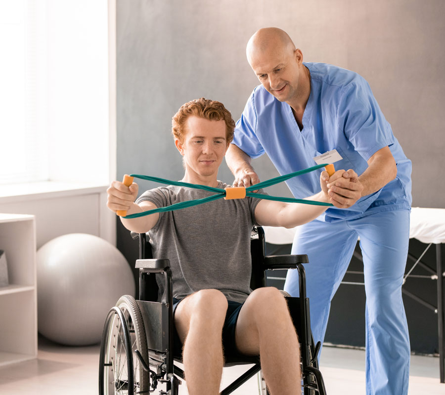 Mature clinician in blue uniform helping young male patient in wheelchair while supporting his left hand during physical exercise with resistance band