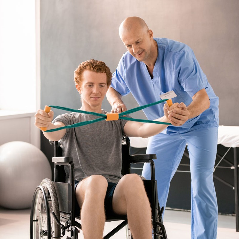 Mature clinician in blue uniform supporting hand of young patient in wheelchair