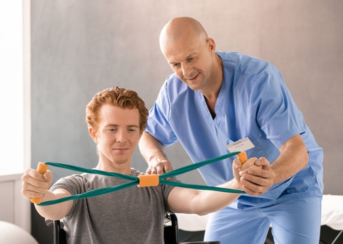Mature clinician in blue uniform supporting hand of young patient in wheelchair